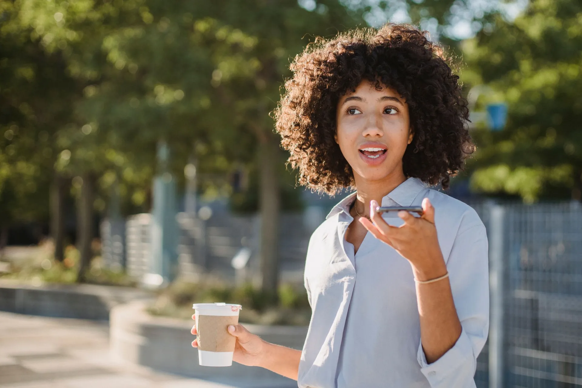 A woman with curly hair talking into her phone outdoors, coffee in hand.