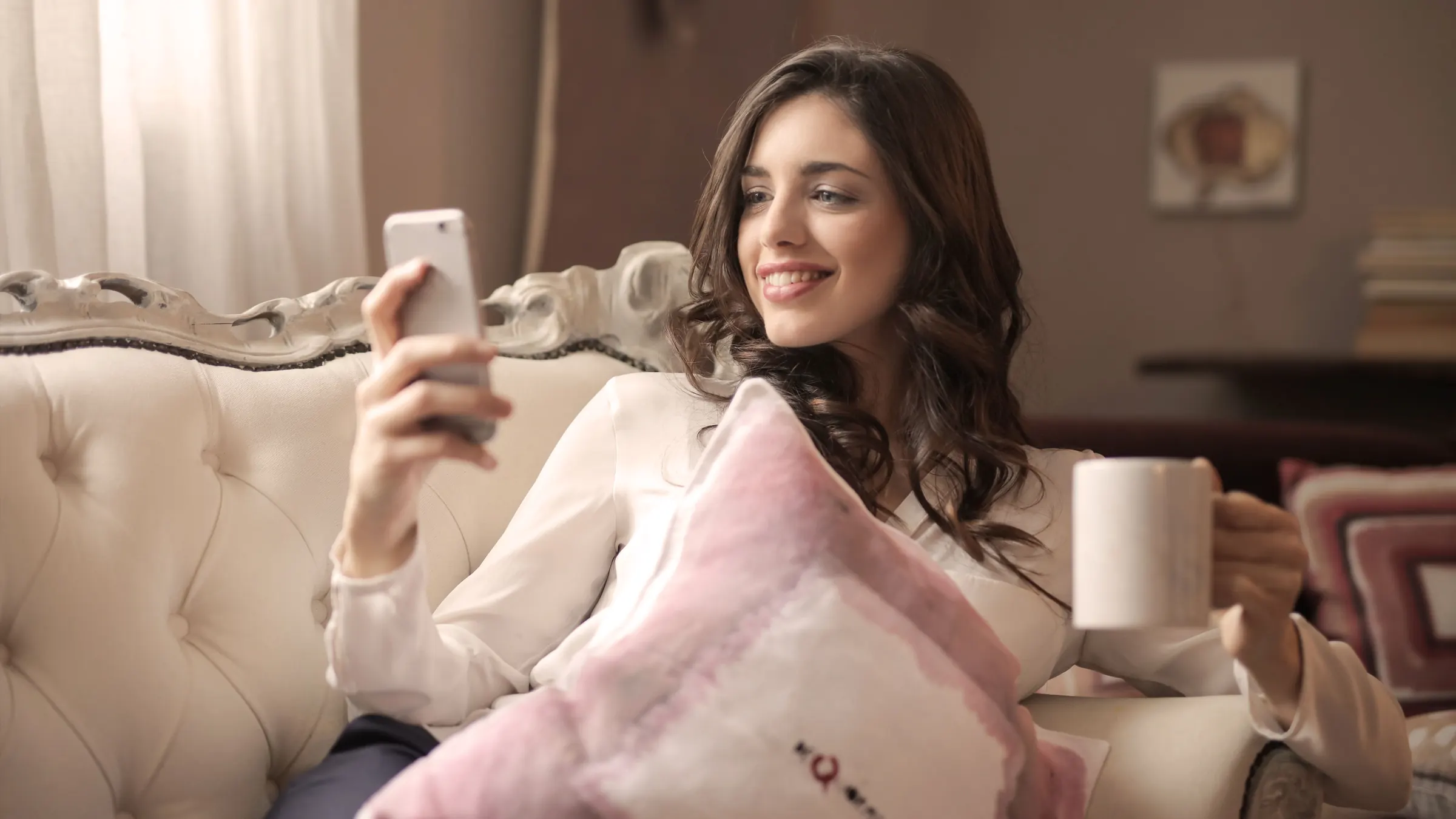 A smiling woman sits on a sofa with a cushion on her lap, looking at her phone and holding a mug.