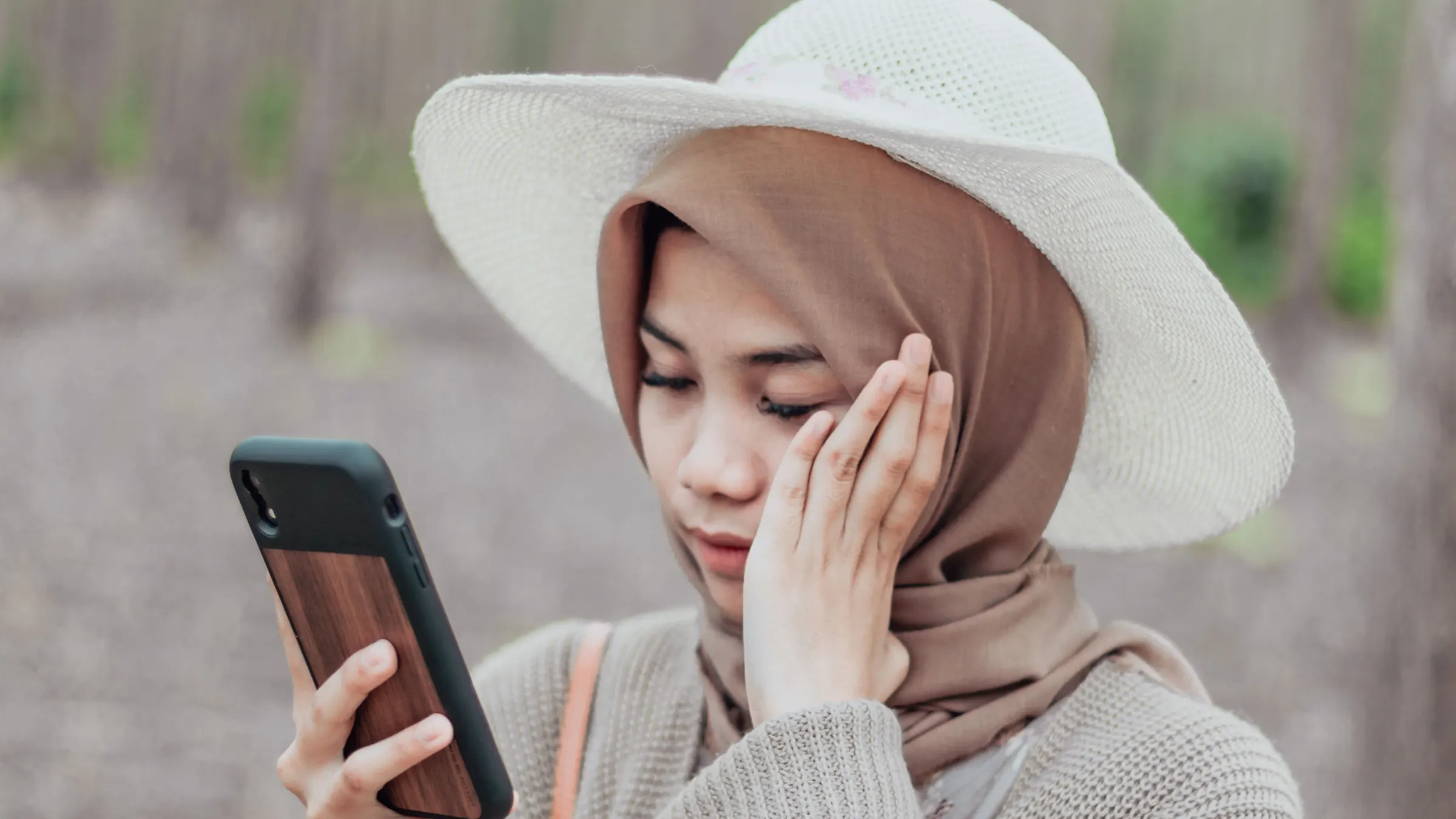 A woman in a wide sun hat and headscarf looks at her phone, one hand resting on her cheek.