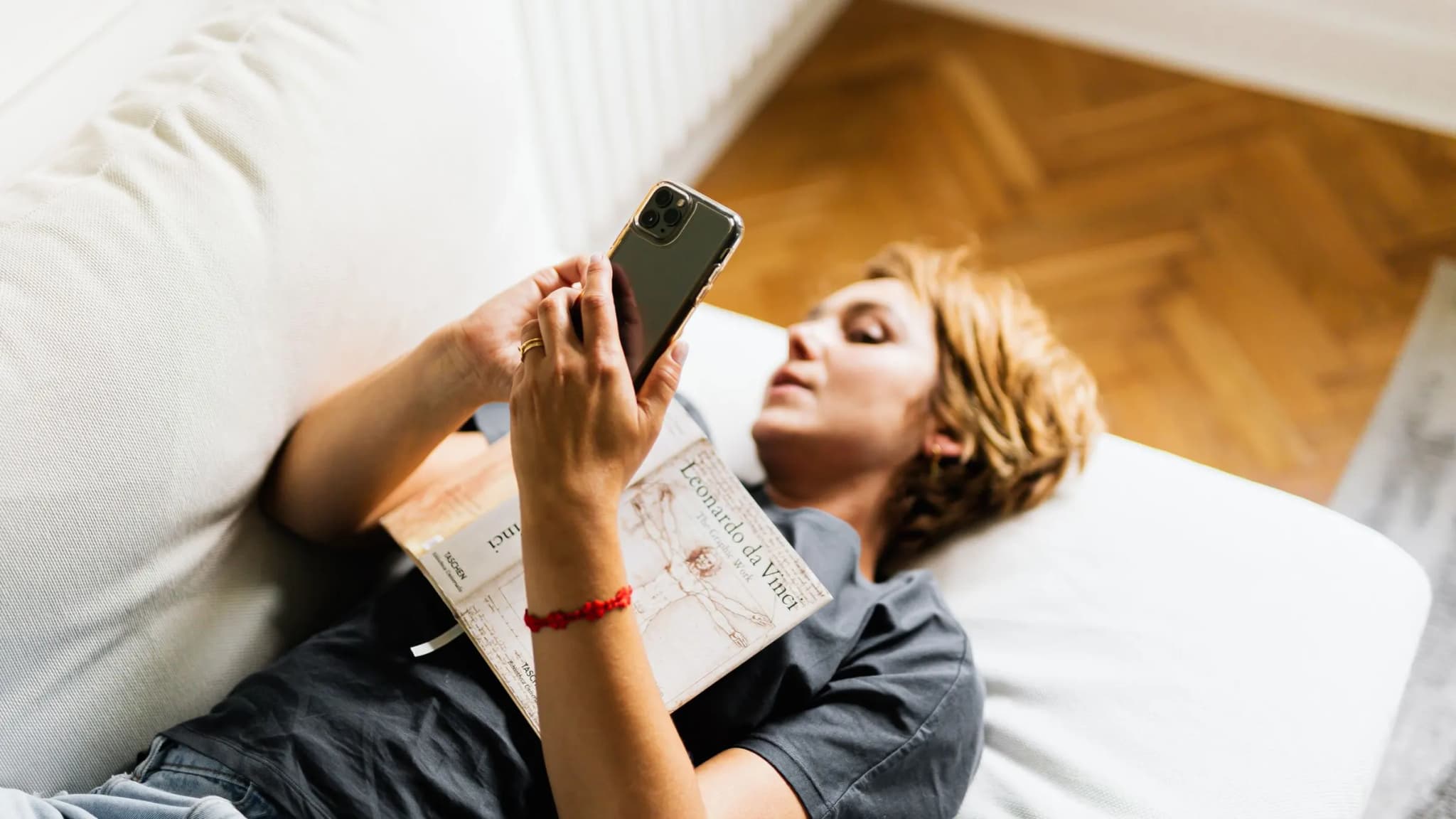 A woman lies on a white sofa, looking at her phone with an open book resting on her chest.