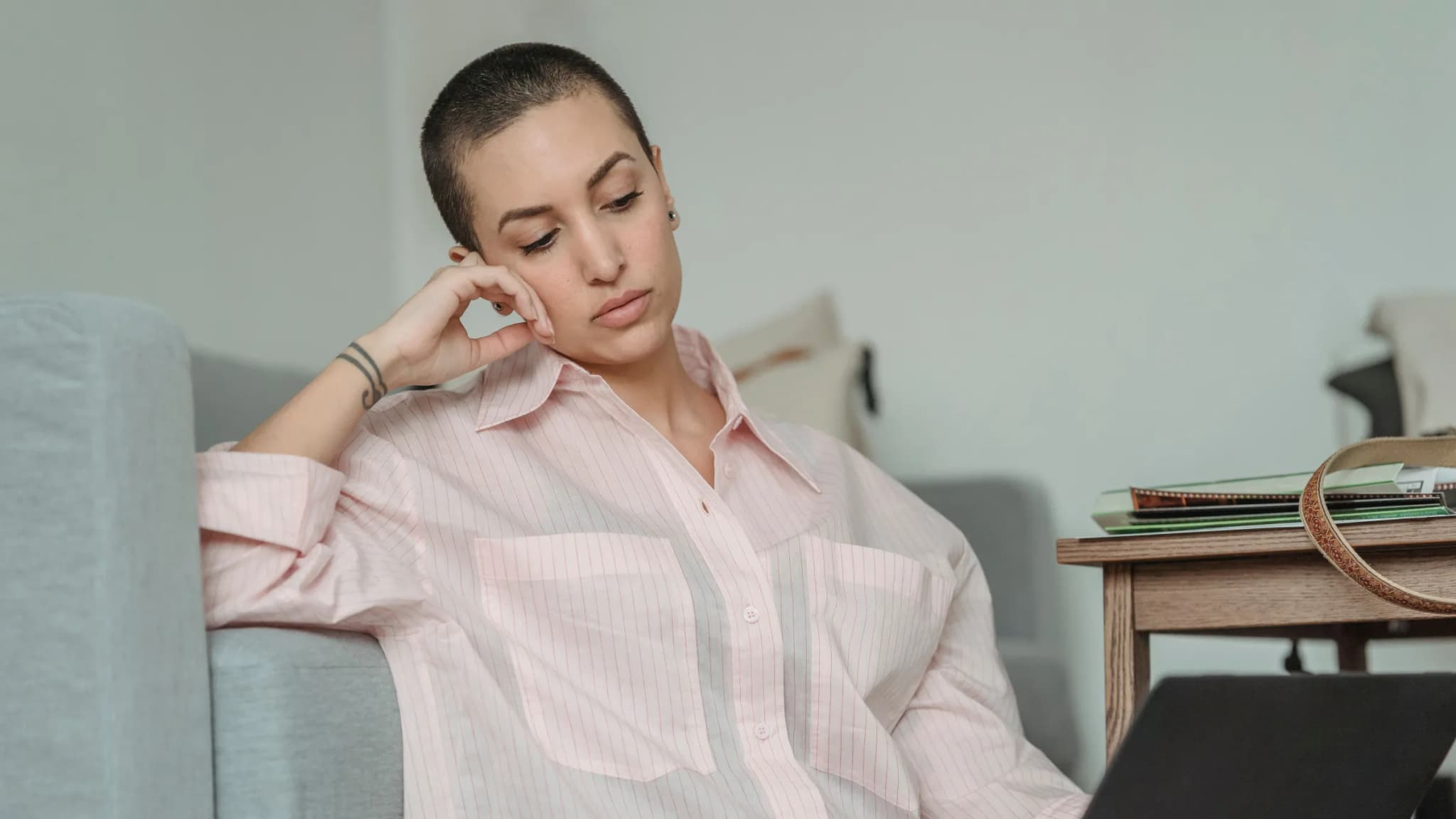 A woman in a pink shirt sits on the floor against a grey sofa, resting her head on her hand and staring at her laptop screen with a tired, unfocused look.