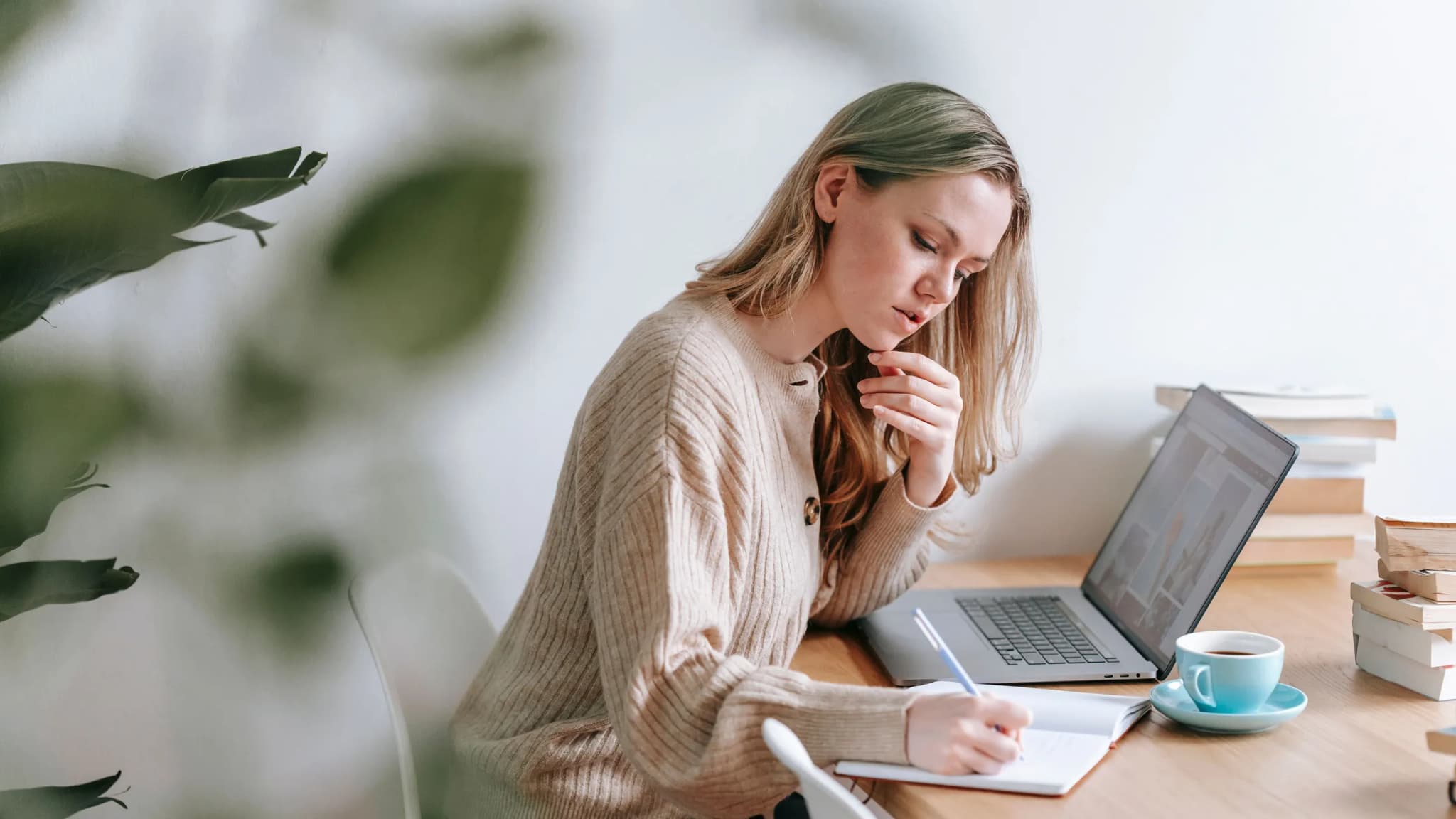 A woman at a desk rests her chin on her hand while writing notes beside a laptop and a cup of coffee