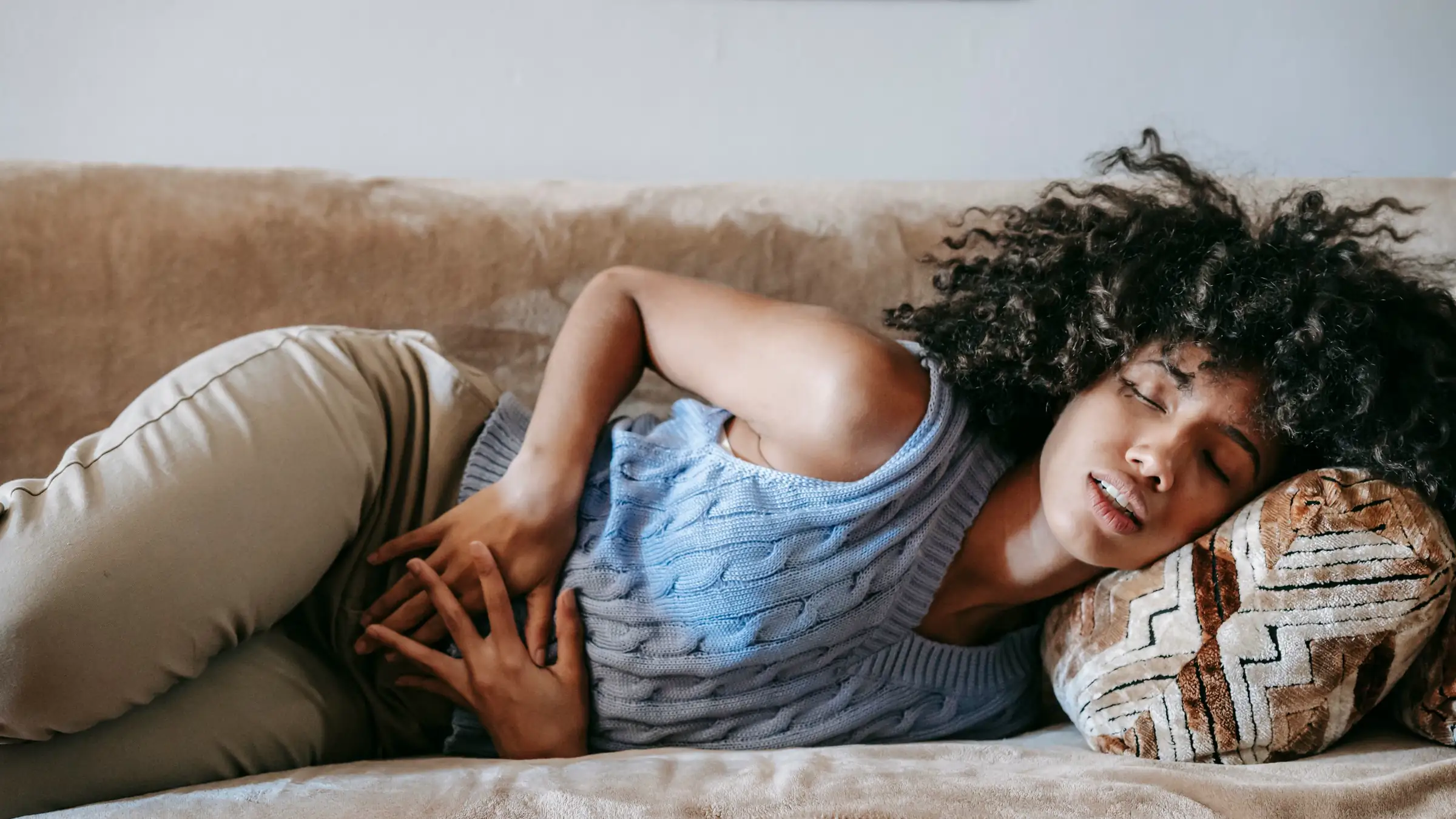 A woman in a blue knitted top lies on her side on a beige sofa with her eyes closed and both hands pressed to her stomach.