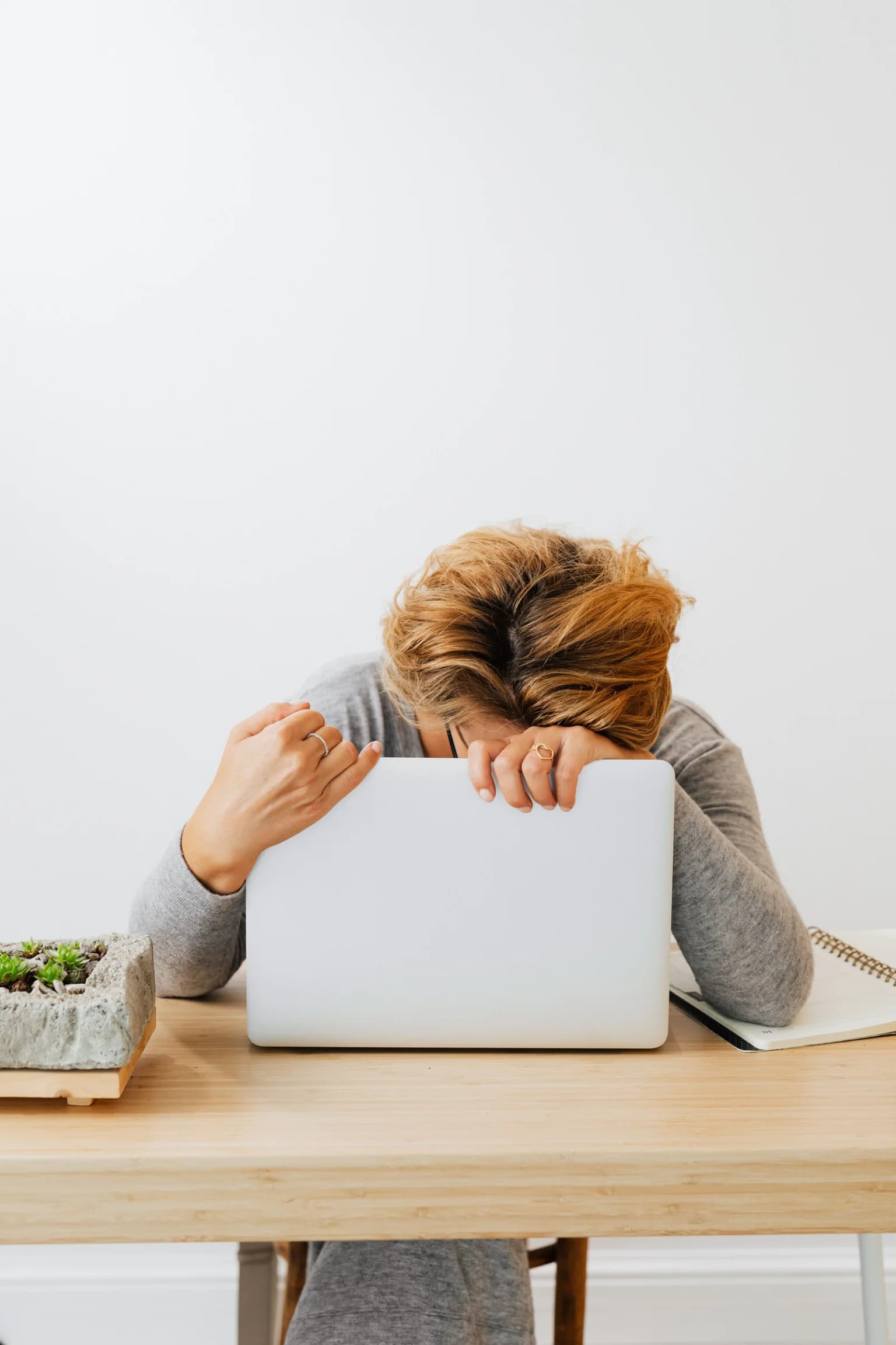 Person leaning forward behind a laptop at a table, resting their head on their hands.