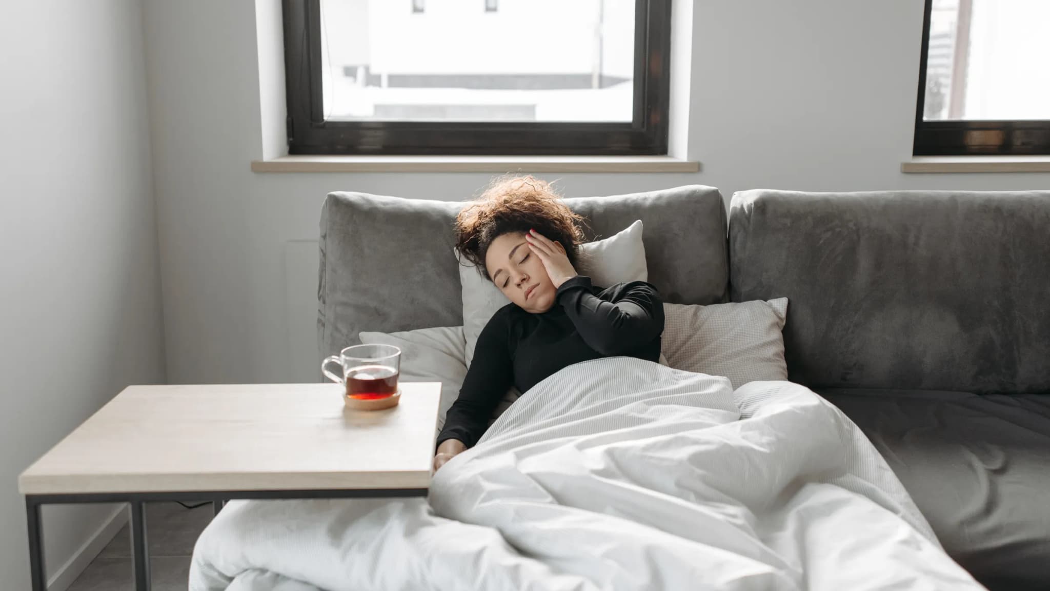 A woman lies back on a grey sofa under a white duvet with her eyes closed and a hand on her head, a glass of tea on the table beside her.