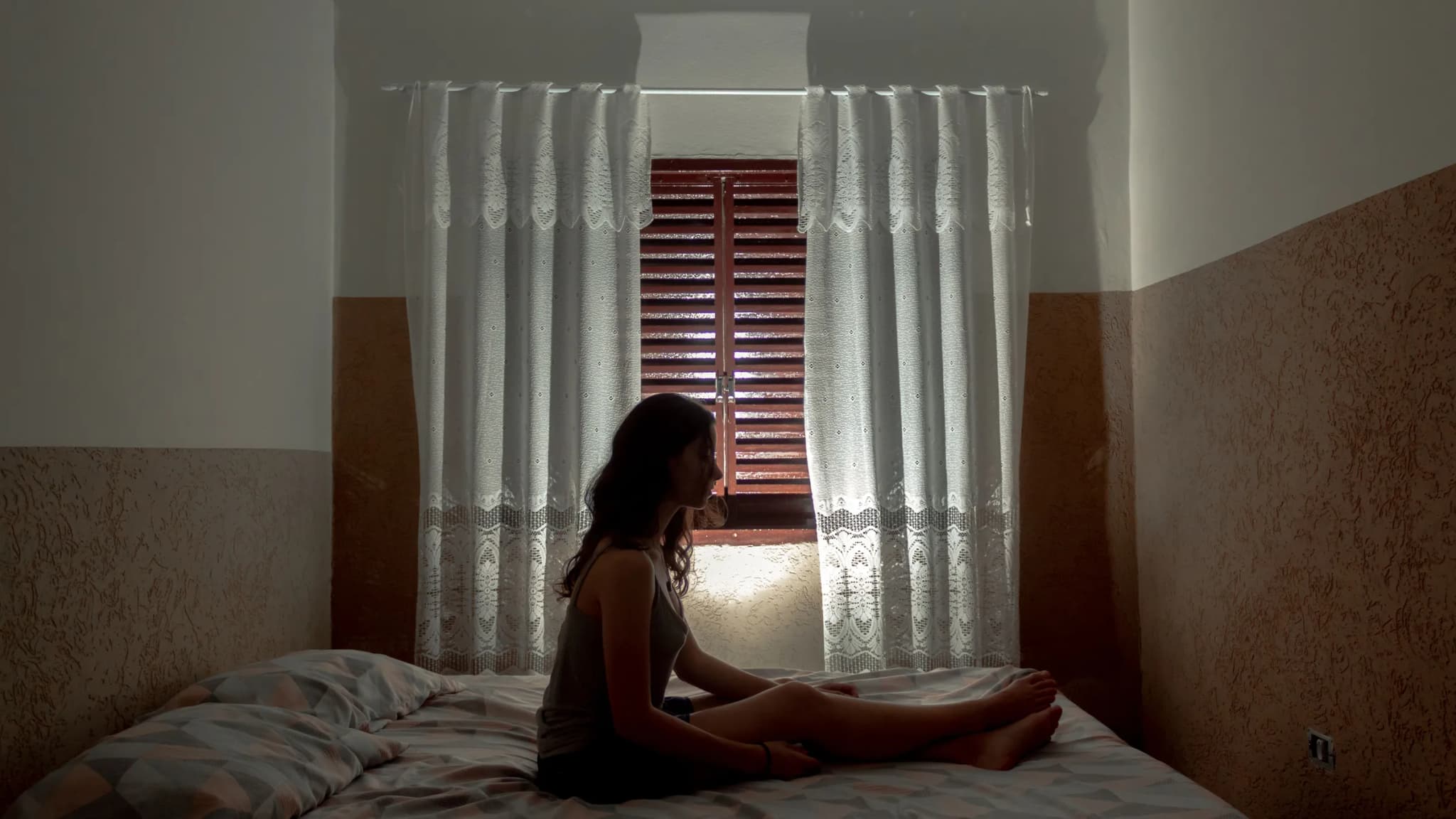 A woman sits on her bed in a dim room, with the shutters closed and lace curtains drawn against the daylight.