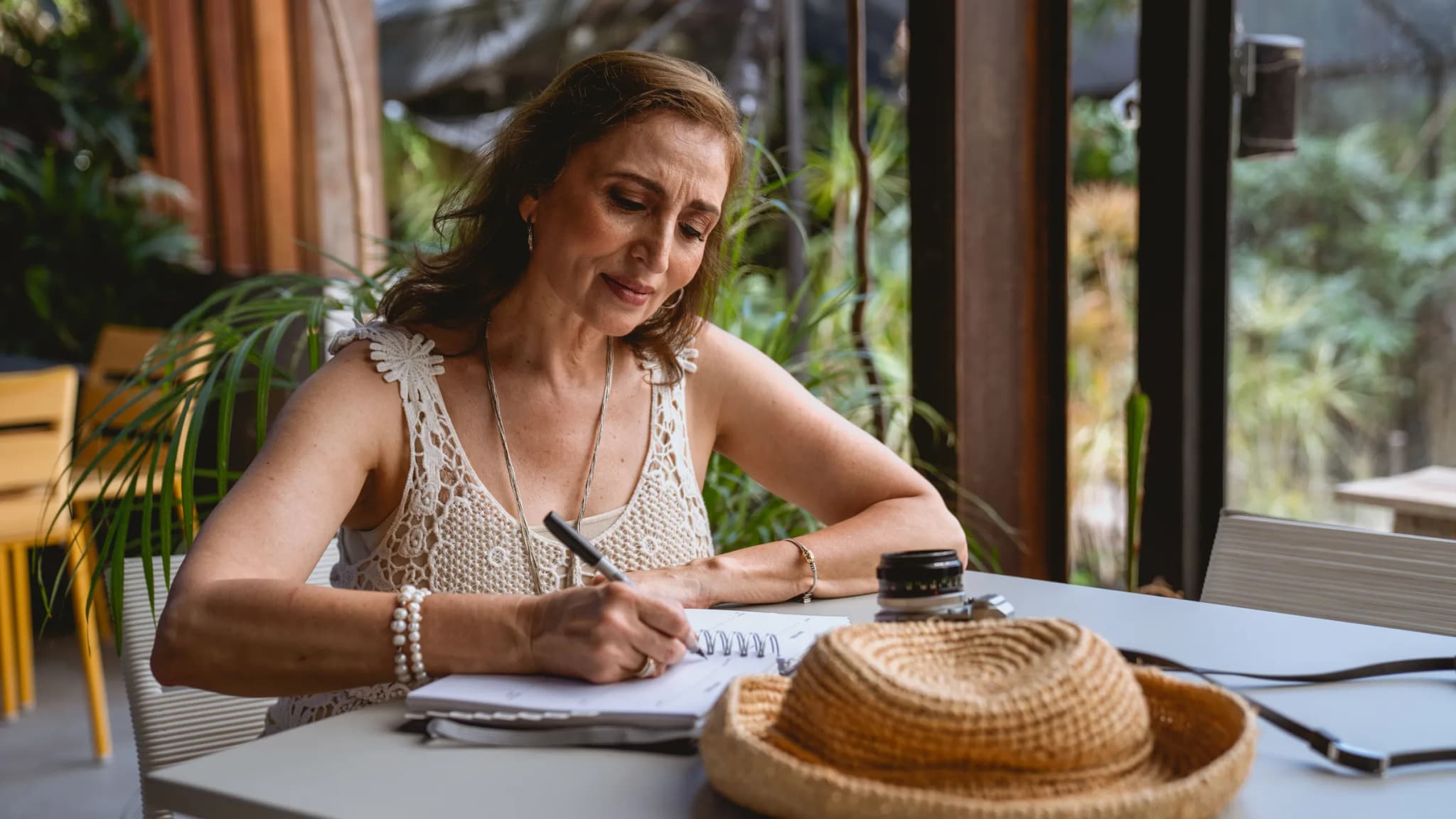 A woman writes in a notebook at a table, with a straw sun hat beside her