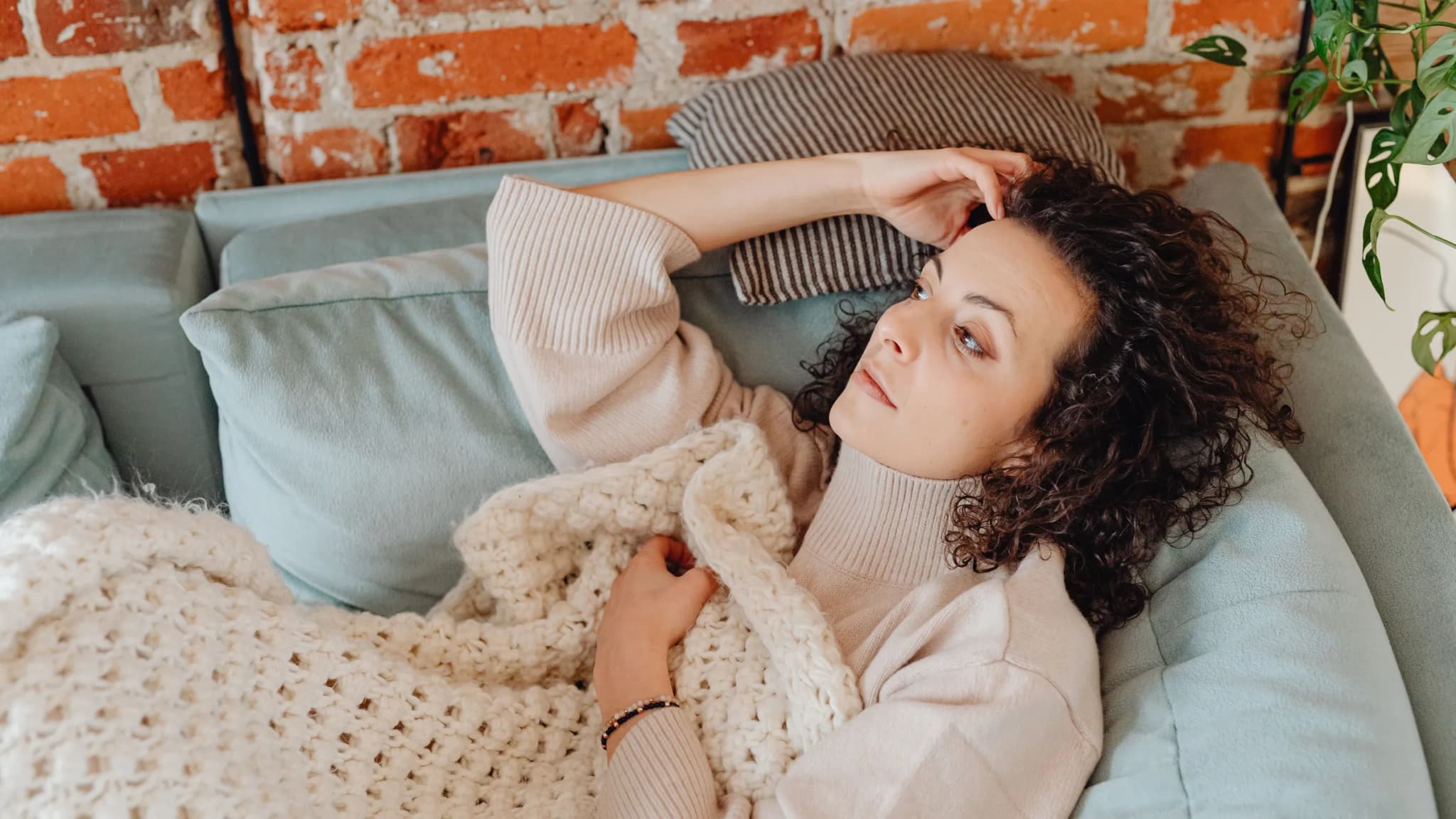 A woman with curly hair rests on a sofa under a knitted blanket.