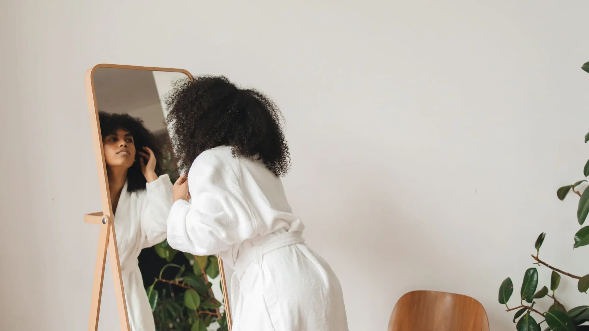 A woman with curly hair in a white bathrobe, seen from behind, leans towards a tall wooden mirror in a bright room with a green plant.