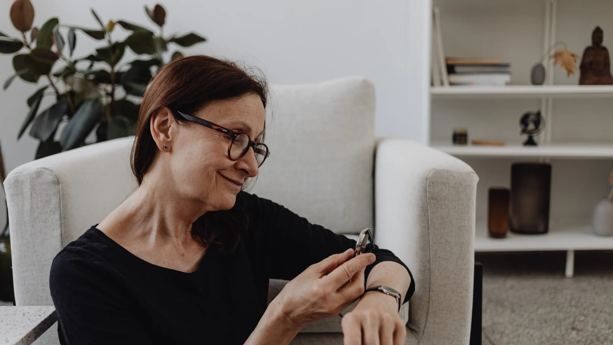 A smiling woman sitting in her living room checks her smartwatch.