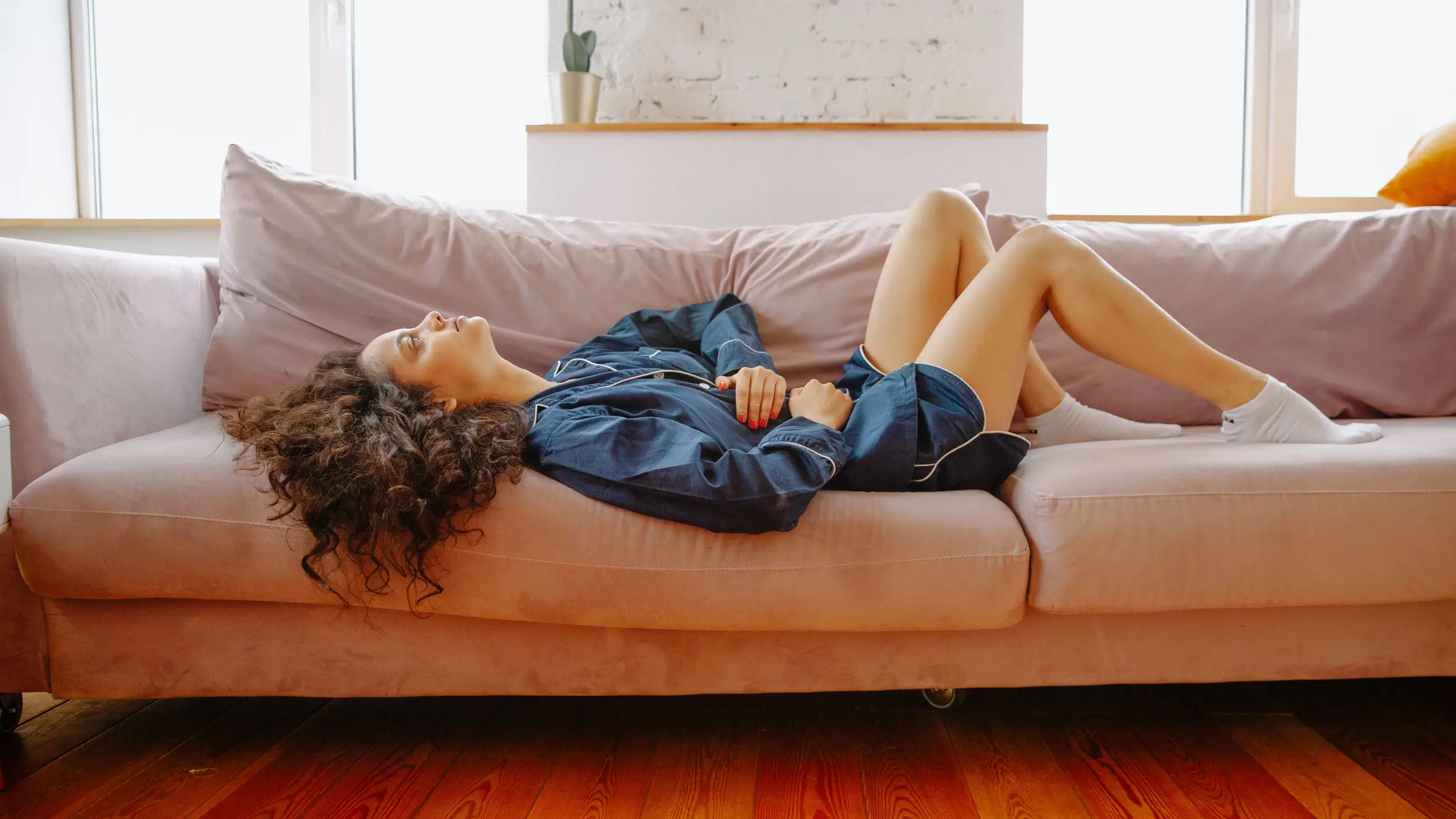 A woman in navy pyjamas lies on her back on a pink sofa with her knees up and both hands resting on her stomach, eyes on the ceiling.