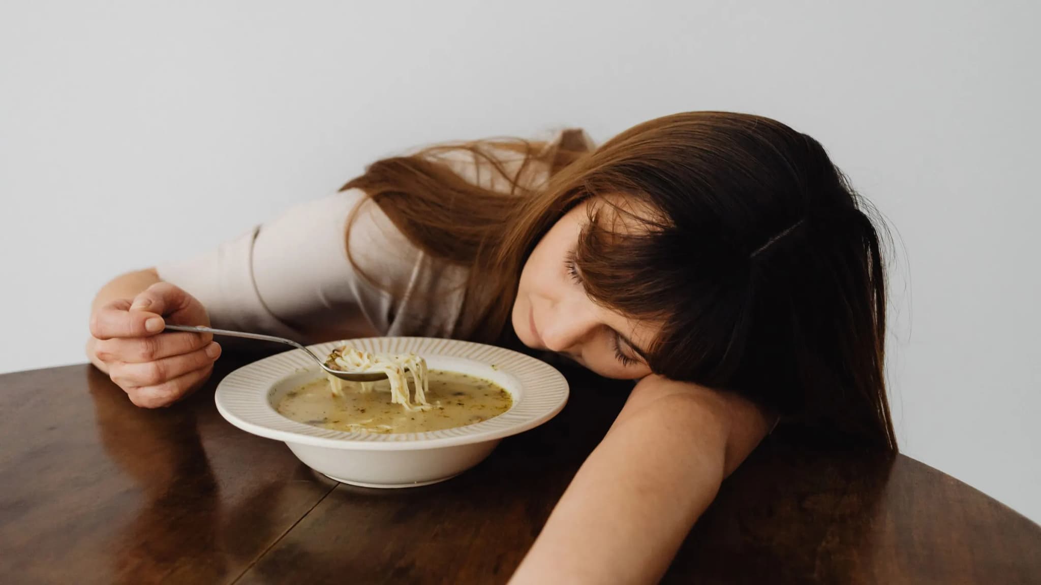 A tired woman rests her head on a table, still holding a spoon over a bowl of soup.