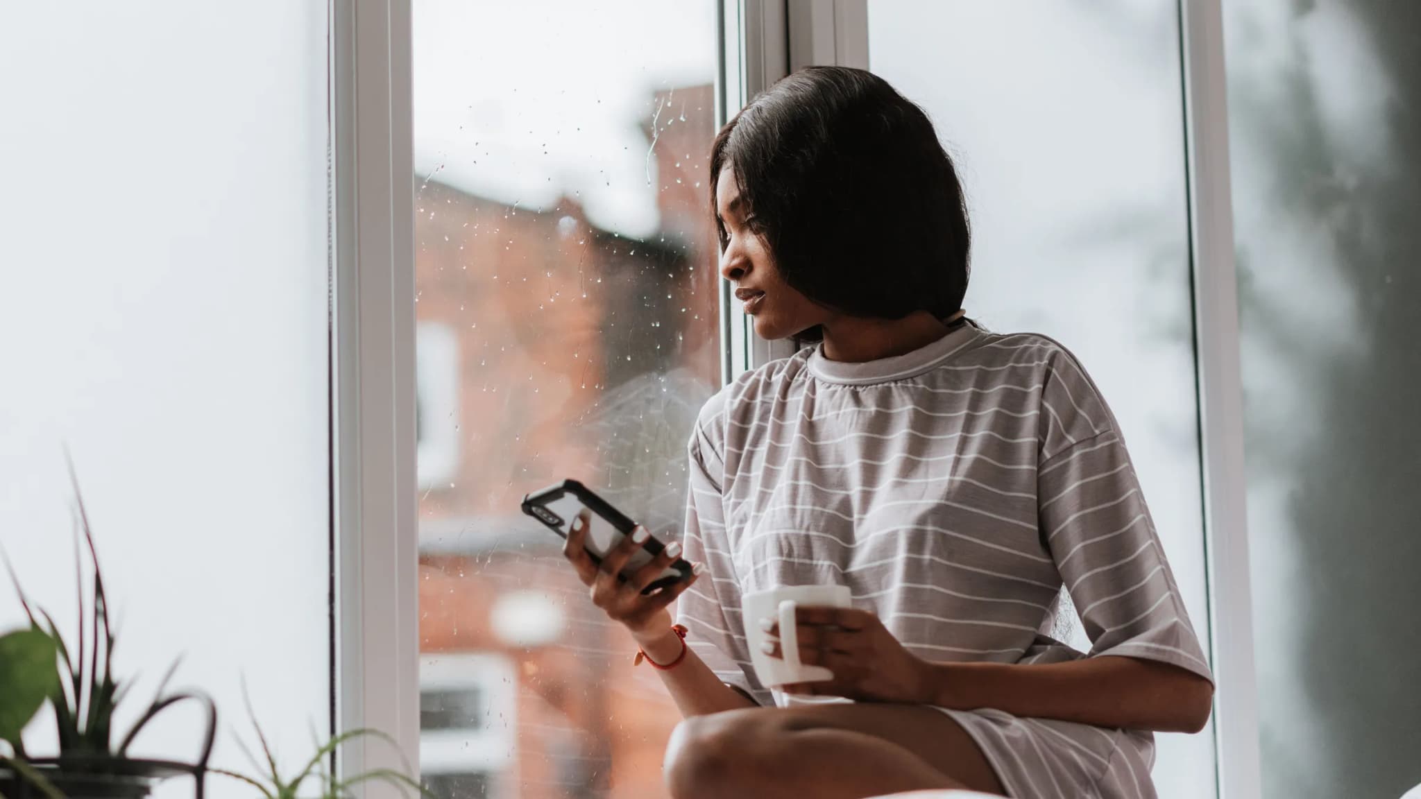 A woman sits by a rain-speckled window holding her phone and a mug.