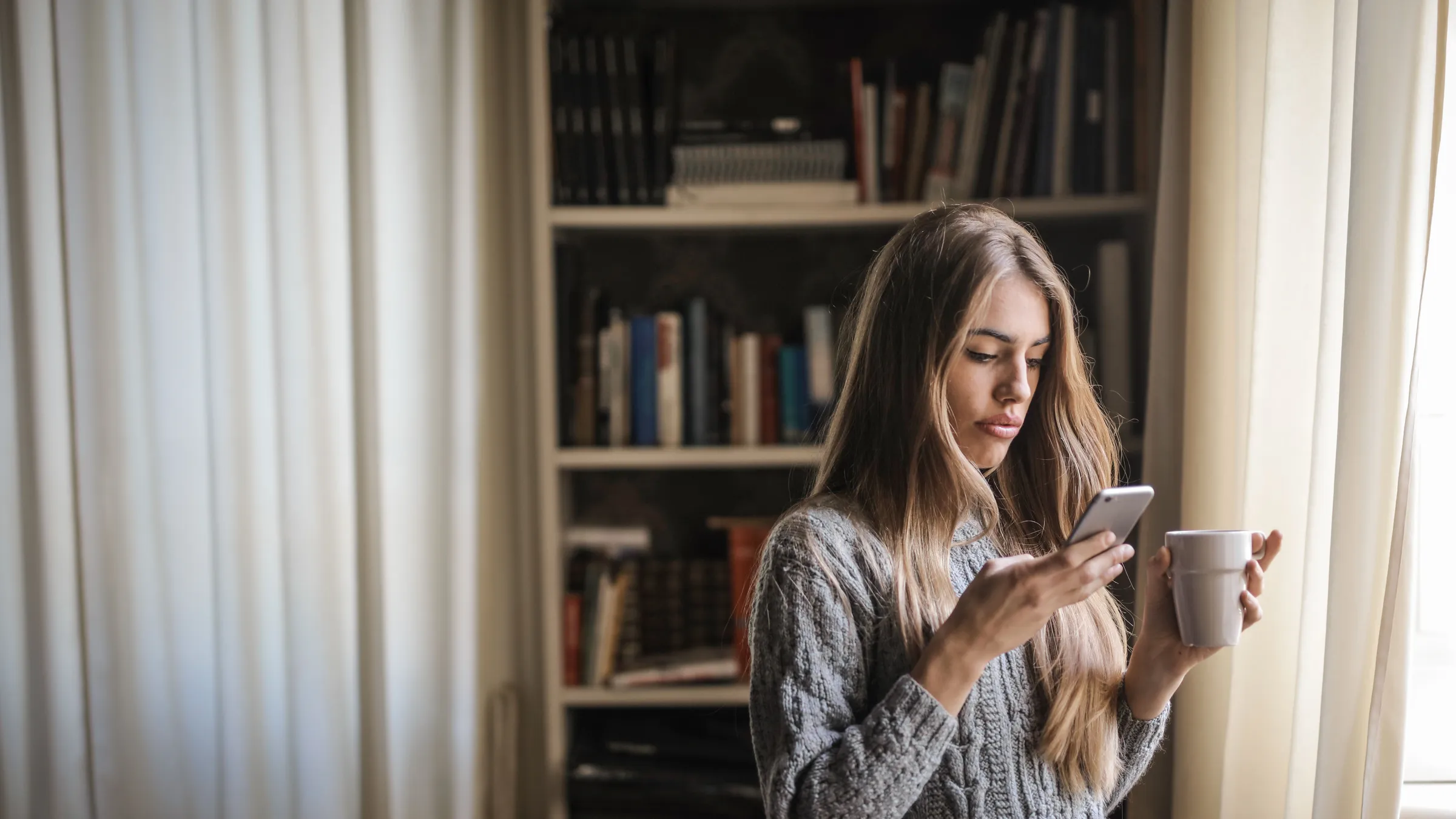 A woman in a grey sweater checks her phone while holding a mug of coffee at home.
