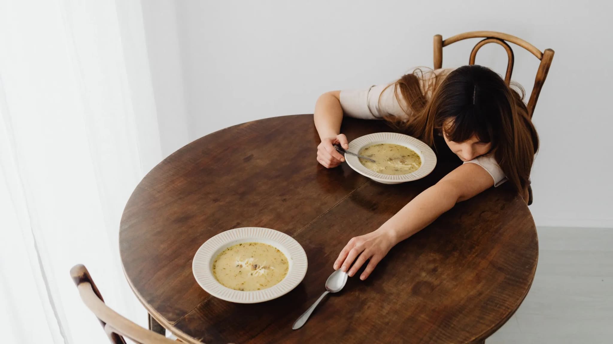 A woman rests her head on a wooden table next to a half-eaten bowl of soup, too tired to finish her meal.