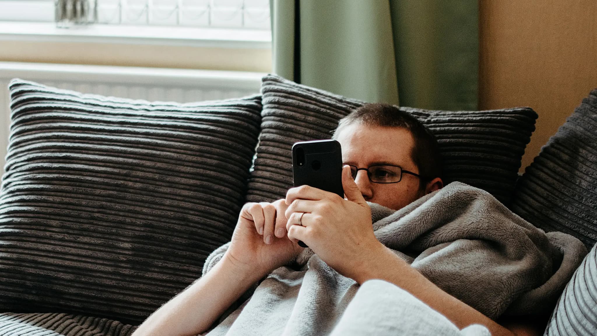 A man lies on a sofa under a blanket, looking at his phone
