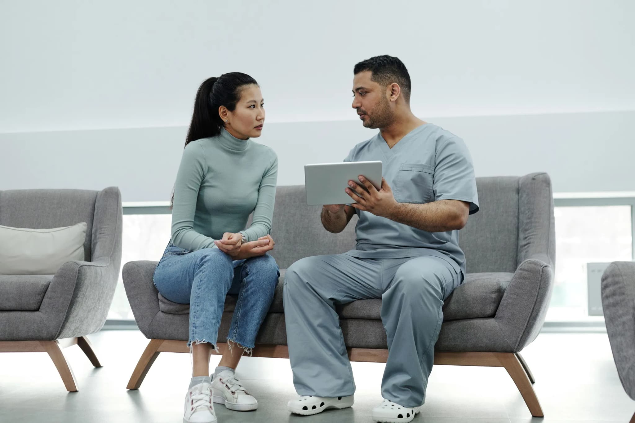 A woman in a green roll-neck top and jeans sits on a grey sofa listening to a clinician in blue scrubs, who holds a tablet.